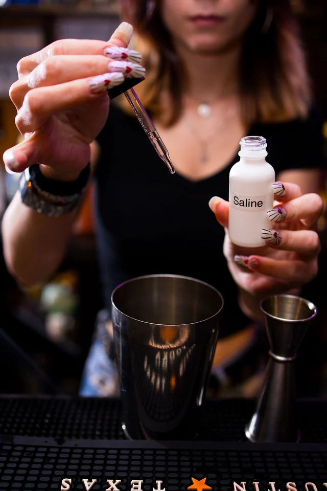 A bartender adds a few drops from a small bottle to a metal shaker.