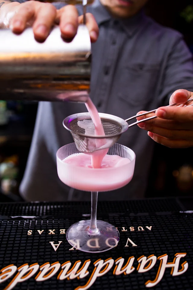 A bartender strains a pink cocktail through a fine strainer into a coupe.