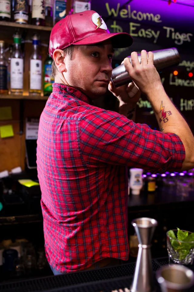 A bartender in a red cap shakes a cocktail near the bar chalkboard.