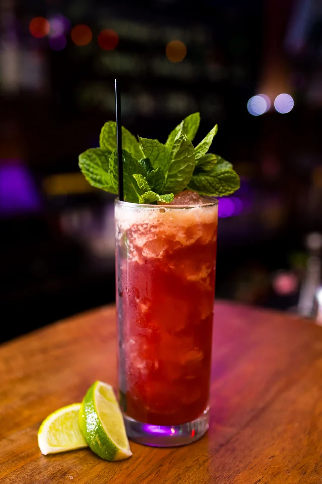 A tall red cocktail with a mint garnish and a cut lime on the bar.