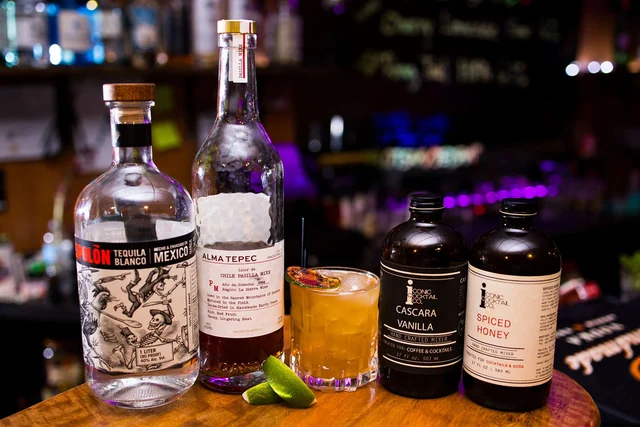 Alma Tepec and bottles labeled Cascara Vanilla and Spiced Honey beside a golden cocktail.